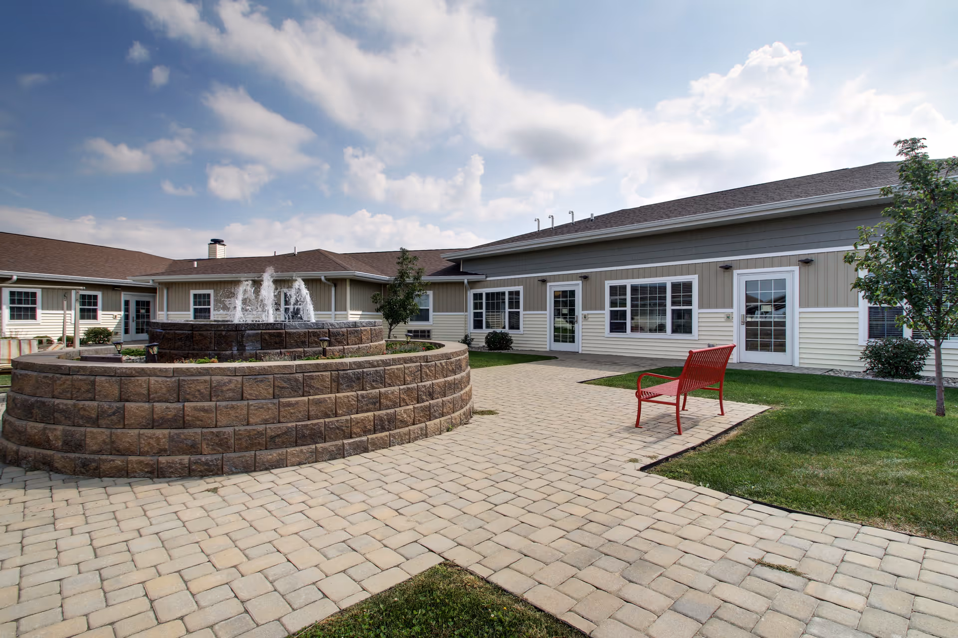 Outdoor courtyard area of a senior care facility with a circular stone water fountain in the center, a paved walkway, a red bench, green grass, small trees, and a beige building with multiple windows and doors under a partly cloudy sky.