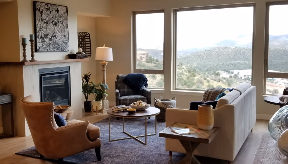 A cozy living room with a large window offering a scenic view of hills and houses. The room features a beige sofa, a brown armchair, a plaid armchair, a round coffee table with decorative items, a side table with a lamp, a fireplace with candles and artwork above it, and several plants.