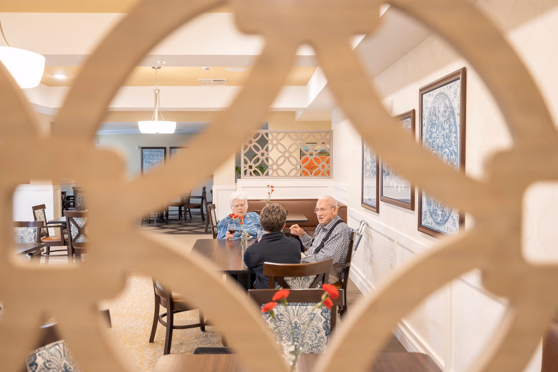 Three elderly people sitting and talking at a dining table in a senior living facility dining room, viewed through a decorative wooden screen. The room has framed artwork on the walls and pendant lighting.