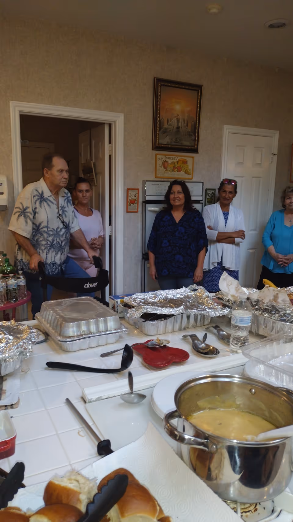 A group of people standing in a kitchen behind a counter covered with foil trays, utensils, a pot of soup, and bread rolls.