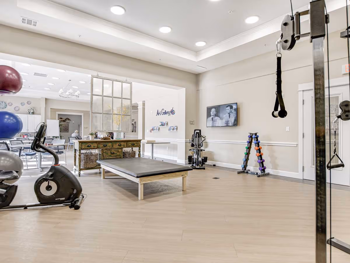A spacious fitness and activity room with exercise equipment including a stationary bike, dumbbells, and resistance bands. There is a large padded bench in the center, a wooden table with baskets underneath, and a wall-mounted TV displaying a black and white image. In the background, there is a dining or activity area with tables and chairs, and a sign on the wall that reads 'Art Gallery'.