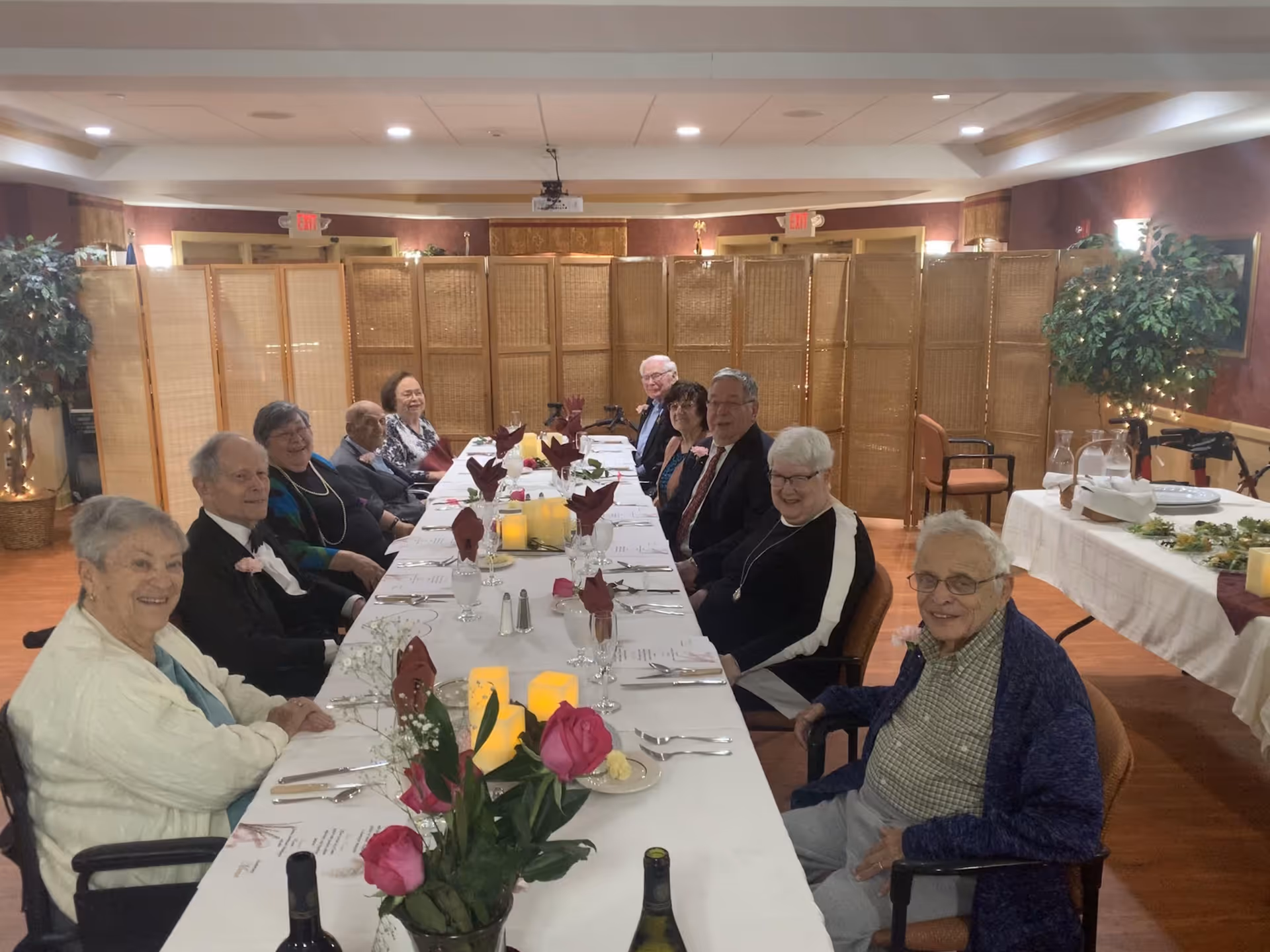 A group of elderly residents seated around a long, formally set dining table in a decorated dining room.