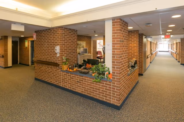 Interior view of a senior living facility hallway with brick walls and a reception desk area decorated with plants and small pumpkins. The hallway is carpeted and has handrails along the walls, with an exit sign visible at the far end.