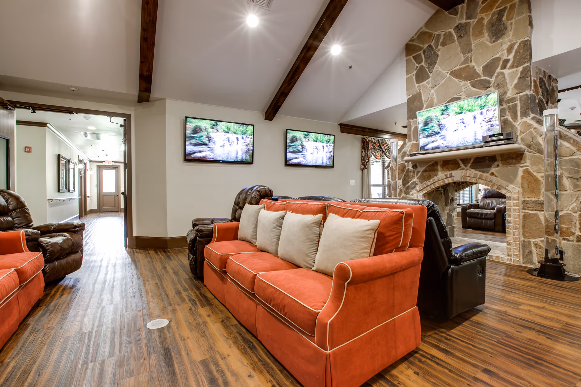 A cozy living room area in Avalon Memory Care featuring two orange sofas with beige pillows, several brown leather recliners, a stone fireplace with a mounted TV above it, and two additional TVs mounted on the wall. The room has wooden flooring and exposed wooden beams on the ceiling.