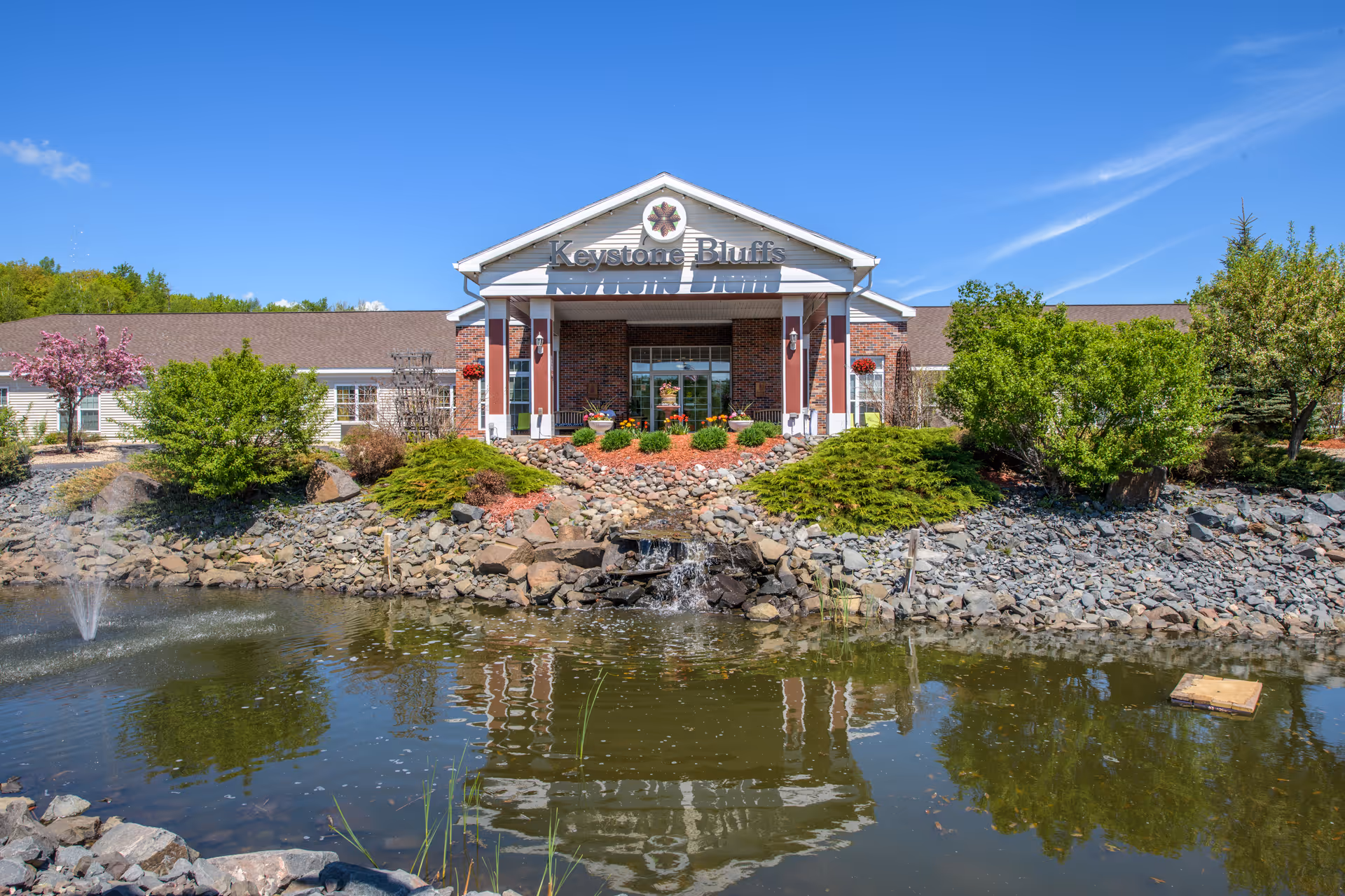 Front exterior view of Keystone Bluffs Assisted Living facility with a pond and small waterfall in the foreground, surrounded by rocks and greenery under a clear blue sky.