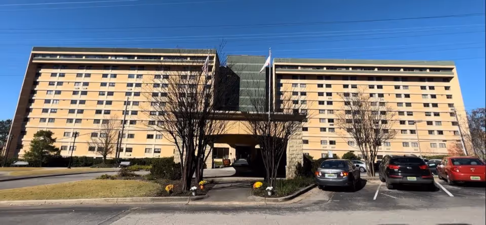 Front exterior of a large multi-story nursing facility with a central covered entrance, flagpoles, trees and a parking lot with cars.