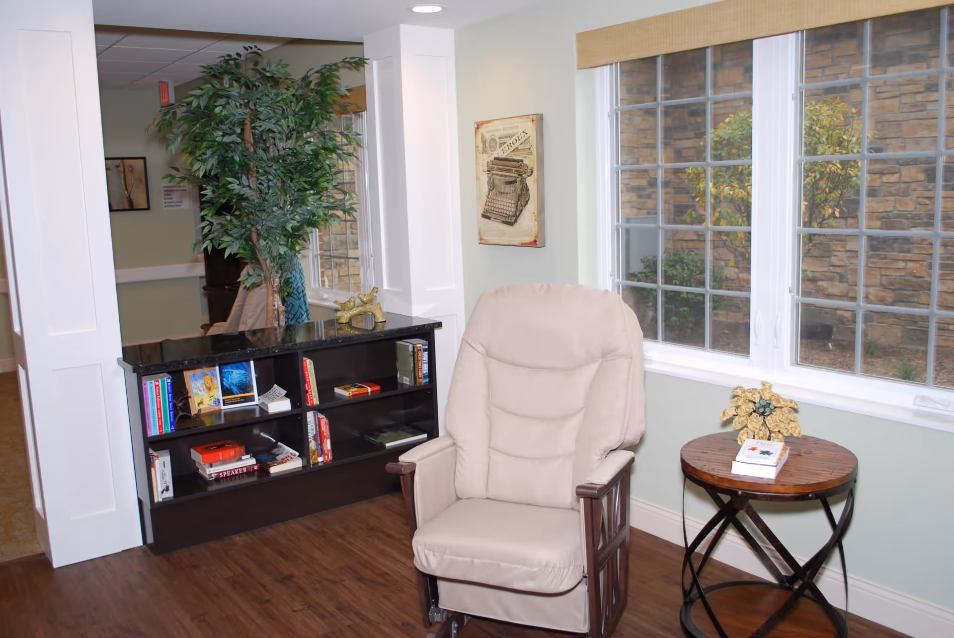 A cozy interior corner of a senior living facility with a beige cushioned armchair next to a round wooden side table holding a small decorative item and a book. Behind the chair is a large window with a view of a stone wall and some greenery outside. To the left, there is a black bookshelf with various books and decorative items, and a tall artificial plant beside it. The floor is wooden, and the walls are painted light green with white trim.