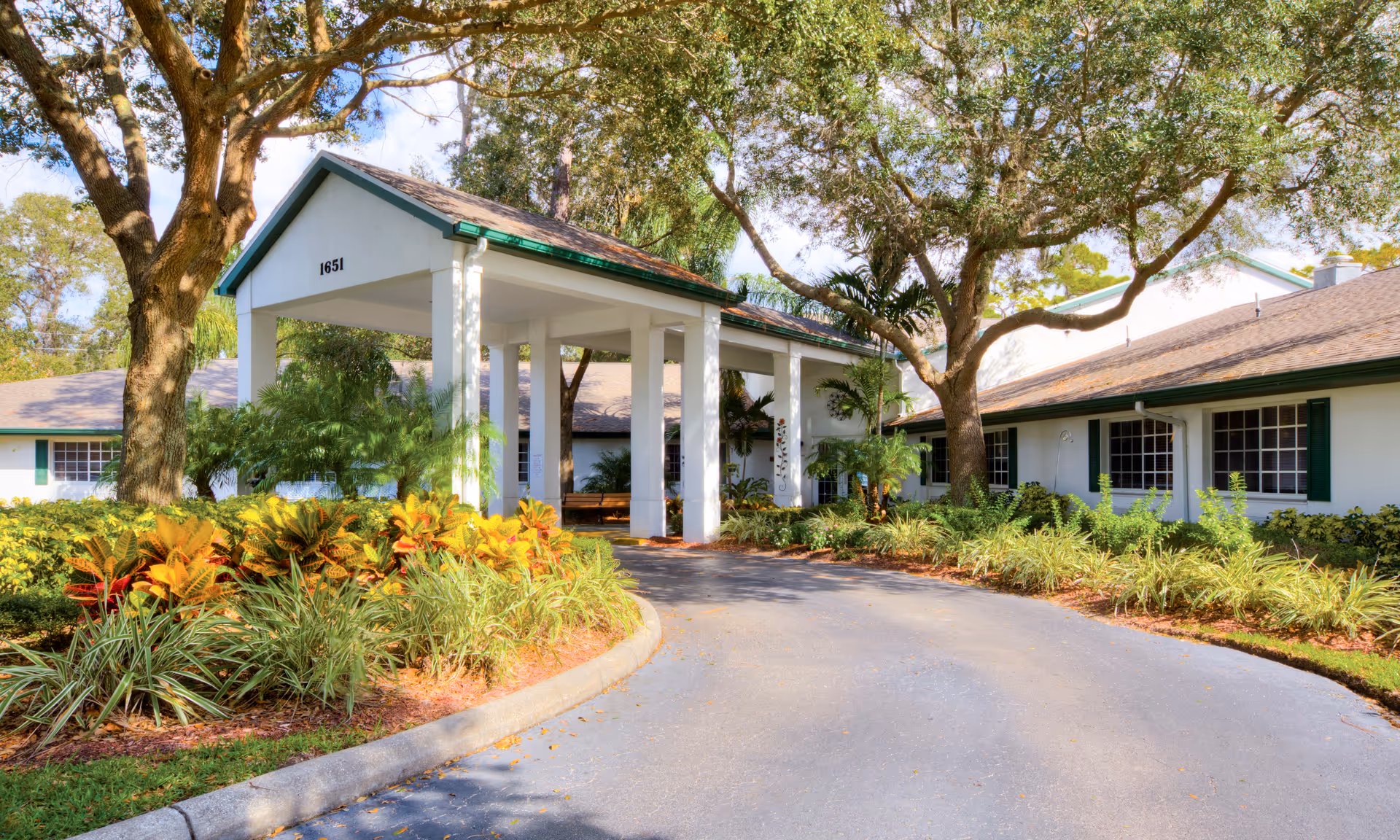 Entrance driveway and covered porte-cochère of a single-story senior living building surrounded by landscaping and trees.