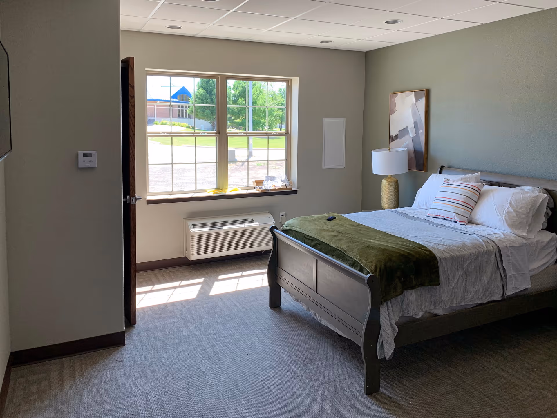 Sunny assisted living bedroom with a neatly made bed, nightstand and large window looking out to greenery.