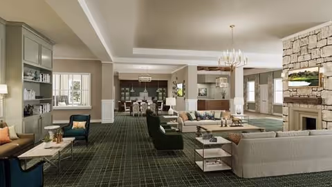 Spacious and elegantly furnished living room area with multiple seating arrangements including sofas and armchairs, a stone fireplace with a mounted TV above it, a chandelier, and large windows allowing natural light to fill the room. The room features a patterned carpet and neutral-colored walls with decorative molding.