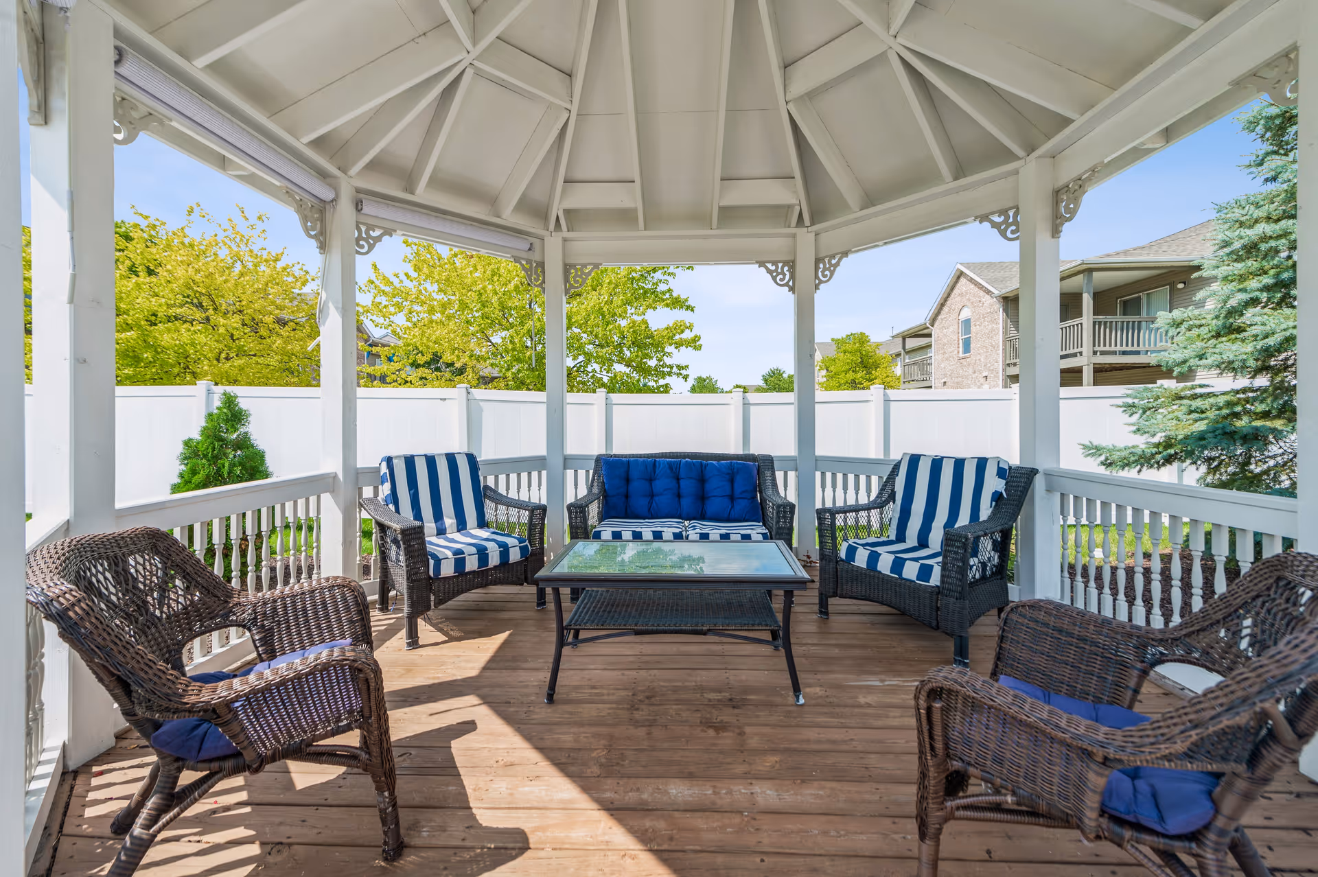 A covered outdoor gazebo with a wooden floor and white railings, furnished with a glass-top coffee table, a blue cushioned loveseat, two striped cushioned armchairs, and two wicker chairs with blue cushions. Green trees and a white fence are visible in the background.