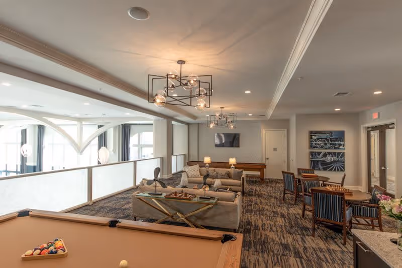 A spacious senior living common area with a pool table in the foreground, comfortable sofas, a coffee table, and several chairs around small tables. The room has modern light fixtures, a television mounted on the wall, and large windows allowing natural light to fill the space.