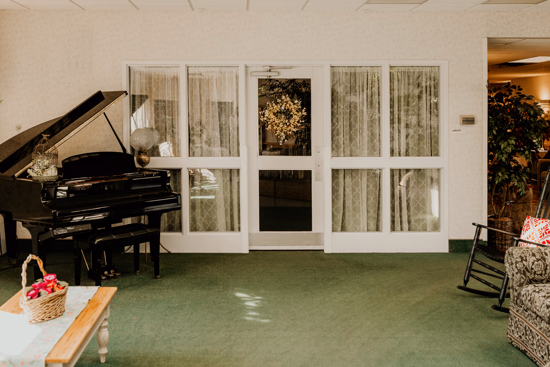 Interior view of a senior living community room featuring a black grand piano on the left, a wooden table with a basket of flowers in the foreground, and a glass door with lace curtains in the center. To the right, there is a rocking chair and a patterned sofa with a red and white pillow. The floor is covered with green carpet and there is a potted plant near the doorway labeled 'Library'.