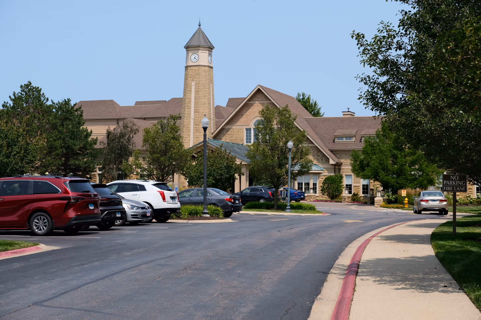 Exterior view of a senior living facility named Vi at The Glen, featuring a large building with a clock tower, surrounded by trees and a parking lot with several parked cars. A sidewalk runs along the right side of the image with a 'No Parking Fire Lane' sign visible.