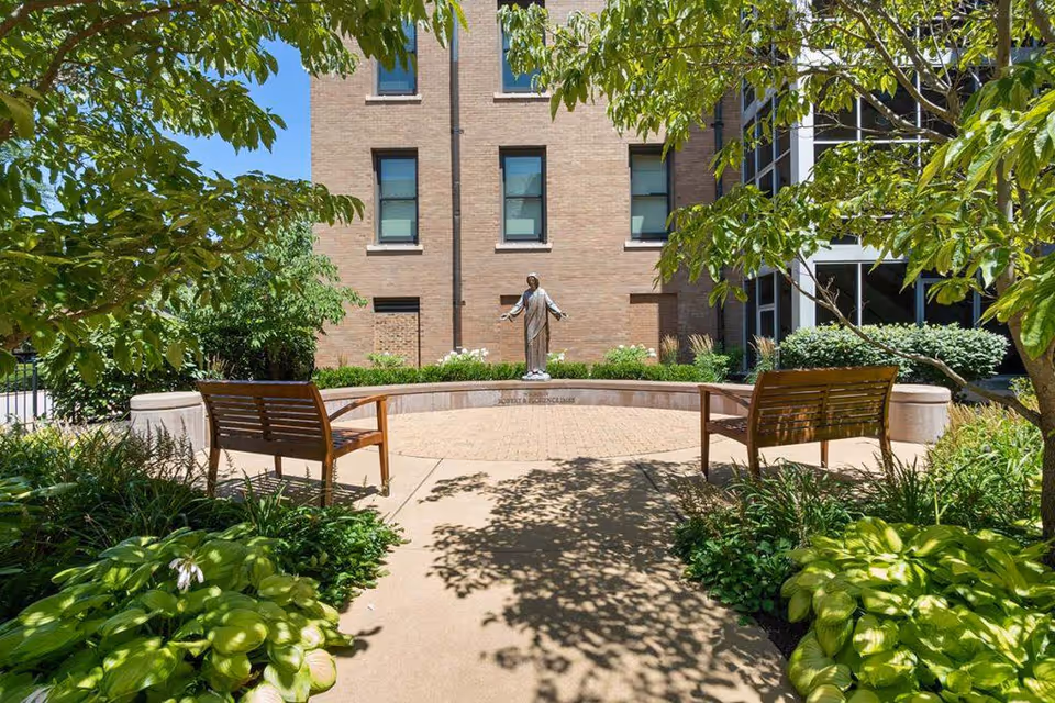 Outdoor courtyard area with two wooden benches facing a statue of a person with outstretched arms, surrounded by green plants and trees, with a brick building in the background under a clear blue sky.