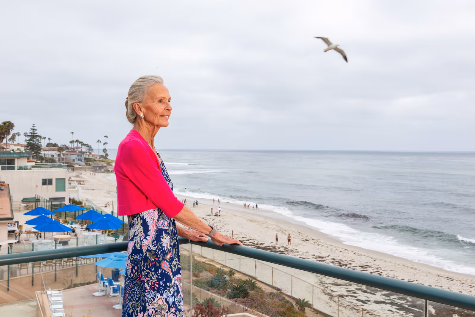 An elderly woman with gray hair tied back, wearing a pink cardigan and a patterned dress, stands on a balcony overlooking a sandy beach and the ocean. Blue umbrellas and outdoor seating are visible below, with people walking along the shoreline and a seagull flying in the cloudy sky.