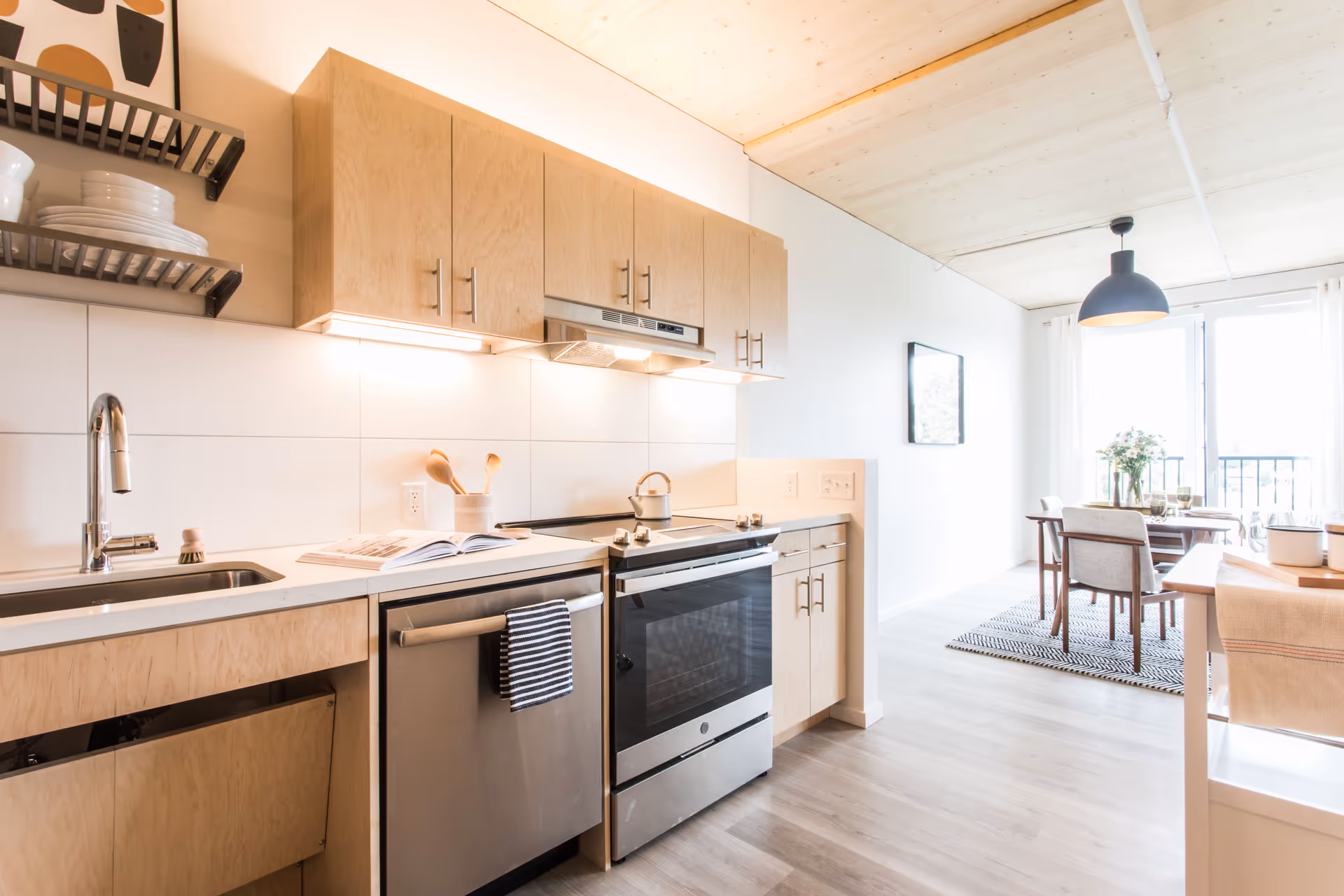 Bright modern kitchen with light wood cabinets, stainless steel dishwasher and oven, white countertops, and a dining area with a table and chairs near a large window with white curtains.