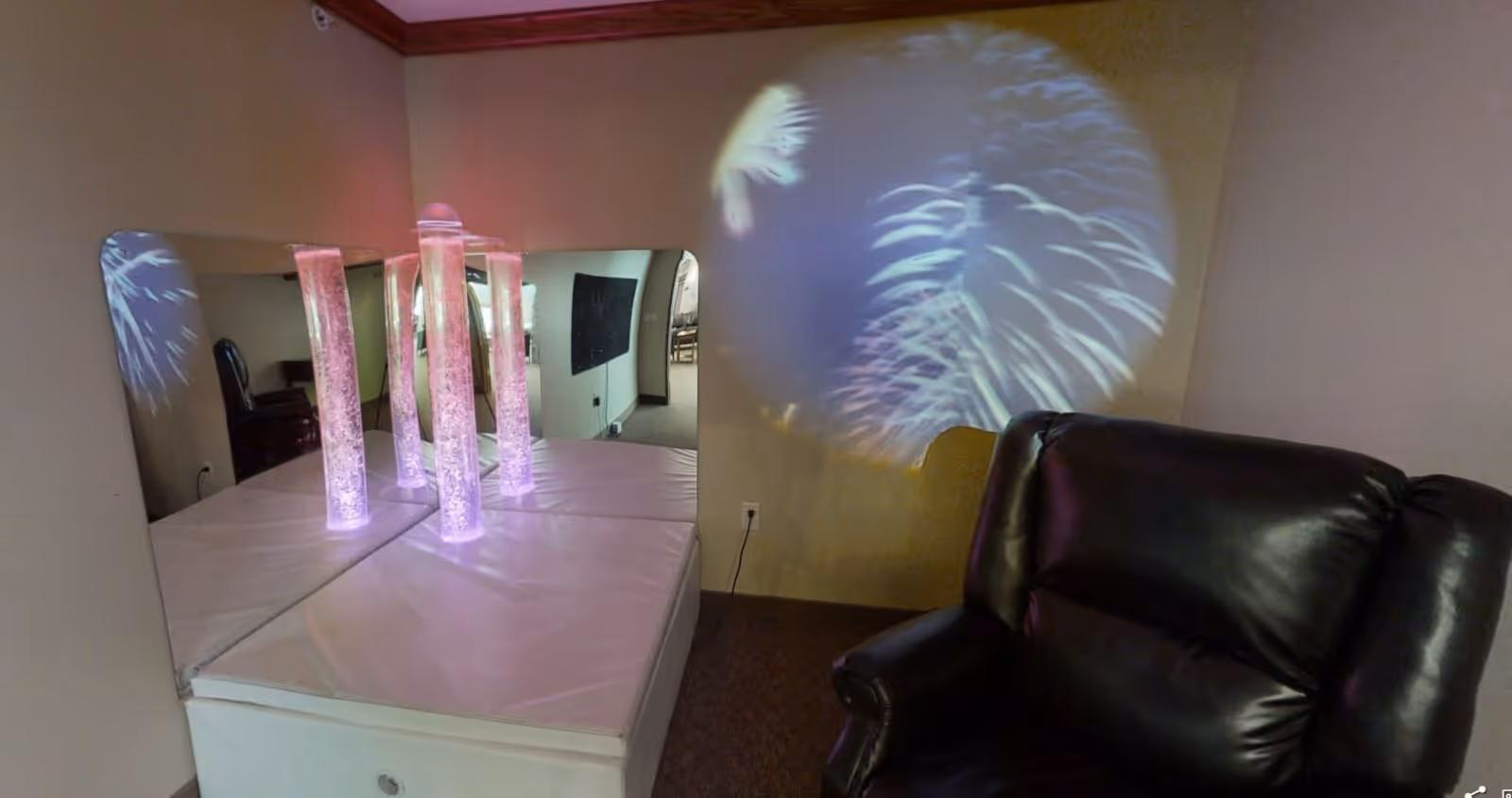 A sensory room corner with a padded platform holding four illuminated bubble tubes emitting soft purple light, a large mirror behind the tubes reflecting the room, a projection of colorful patterns on the wall, and a black leather recliner chair nearby.