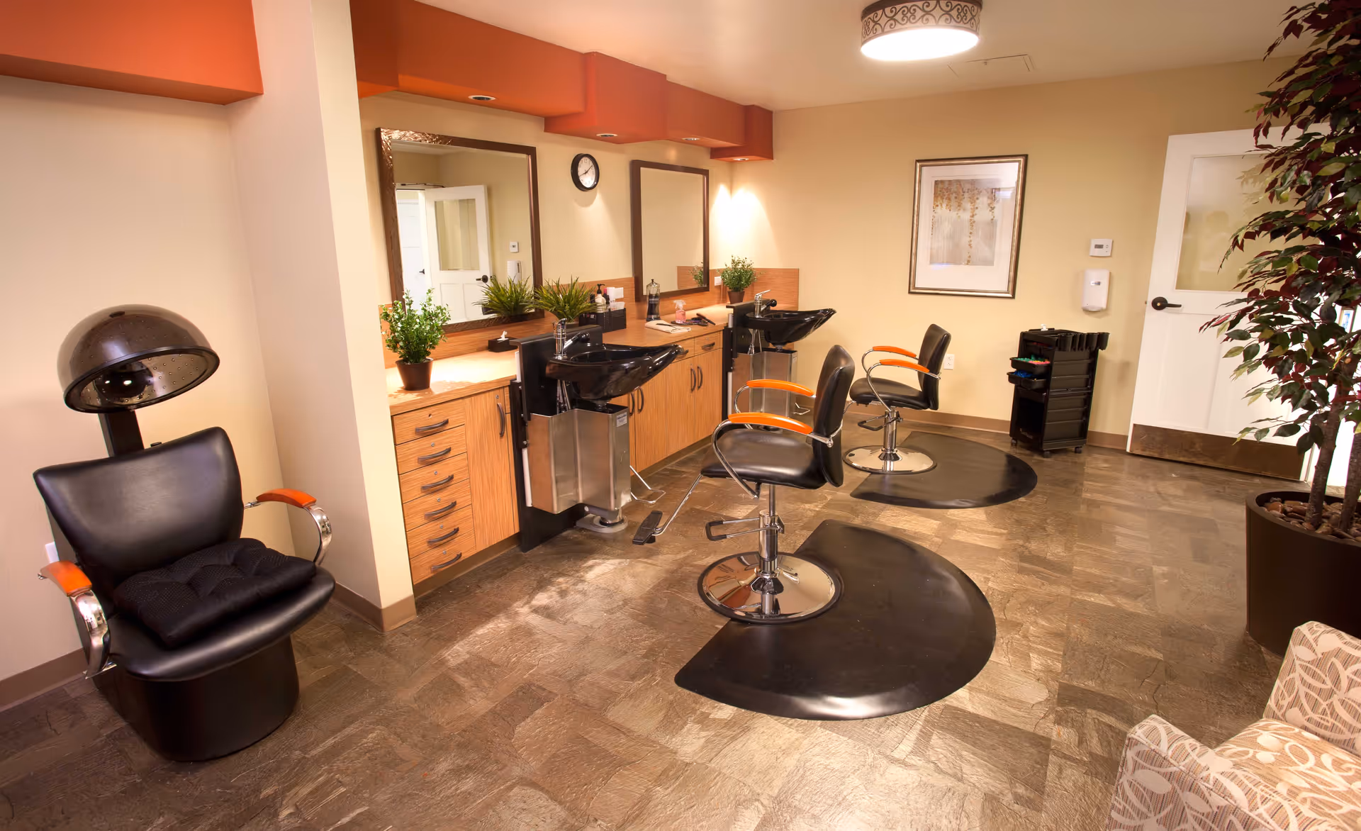 Bright salon-style interior with styling chairs, hair-washing sinks, mirrors and a hood dryer.