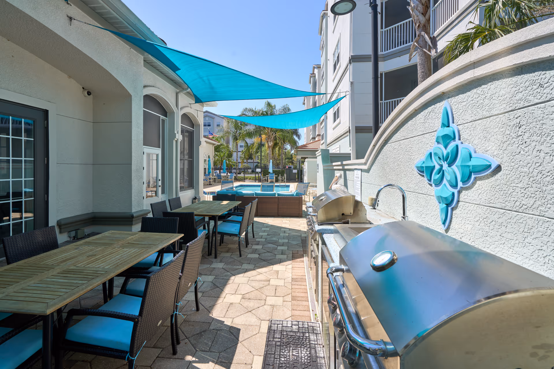 Outdoor patio area with wooden tables and wicker chairs with blue cushions, a stainless steel barbecue grill, and blue shade sails overhead. In the background, there is a swimming pool surrounded by palm trees and additional seating.