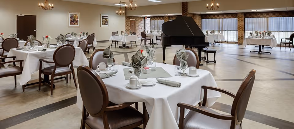 A spacious dining room with multiple round tables covered in white tablecloths, each set with folded napkins, cups, glasses, and small flower vases. The room features large windows with sheer curtains, brick pillars, and a black grand piano near the center. The walls are decorated with framed artwork, and chandeliers hang from the ceiling providing warm lighting.