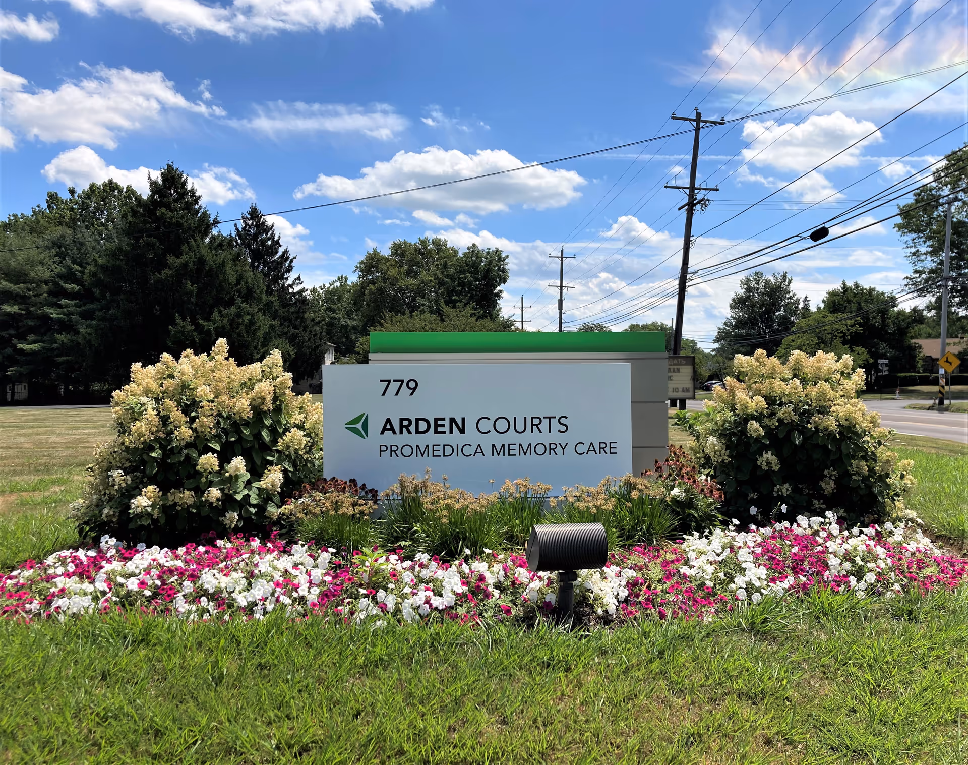 Outdoor view of a sign for Arden Courts ProMedica Memory Care Community at 779, surrounded by colorful flowers and greenery under a partly cloudy blue sky.