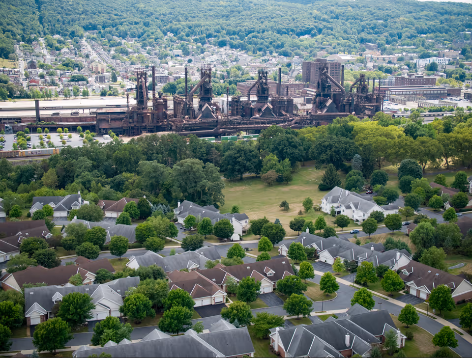 Aerial view of a residential neighborhood with many houses surrounded by trees and greenery. In the background, there is a large industrial complex with tall structures and smokestacks. Beyond the industrial area, a town with numerous buildings and a forested hillside is visible.