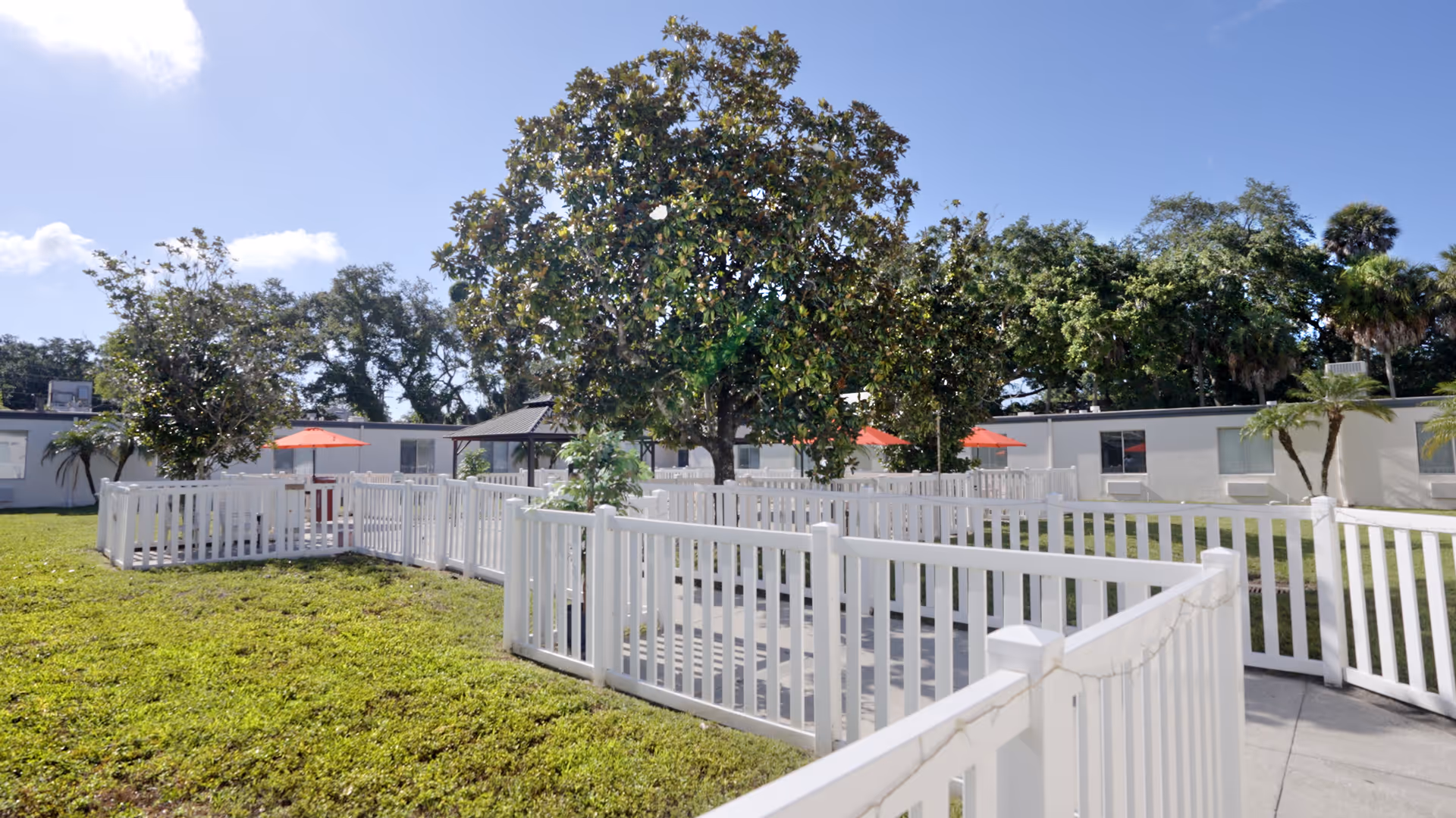 Outdoor area of Woodbridge Care Center featuring a white picket fence, green grass, several trees, and a few red umbrellas providing shade over seating areas. The sky is clear and blue.