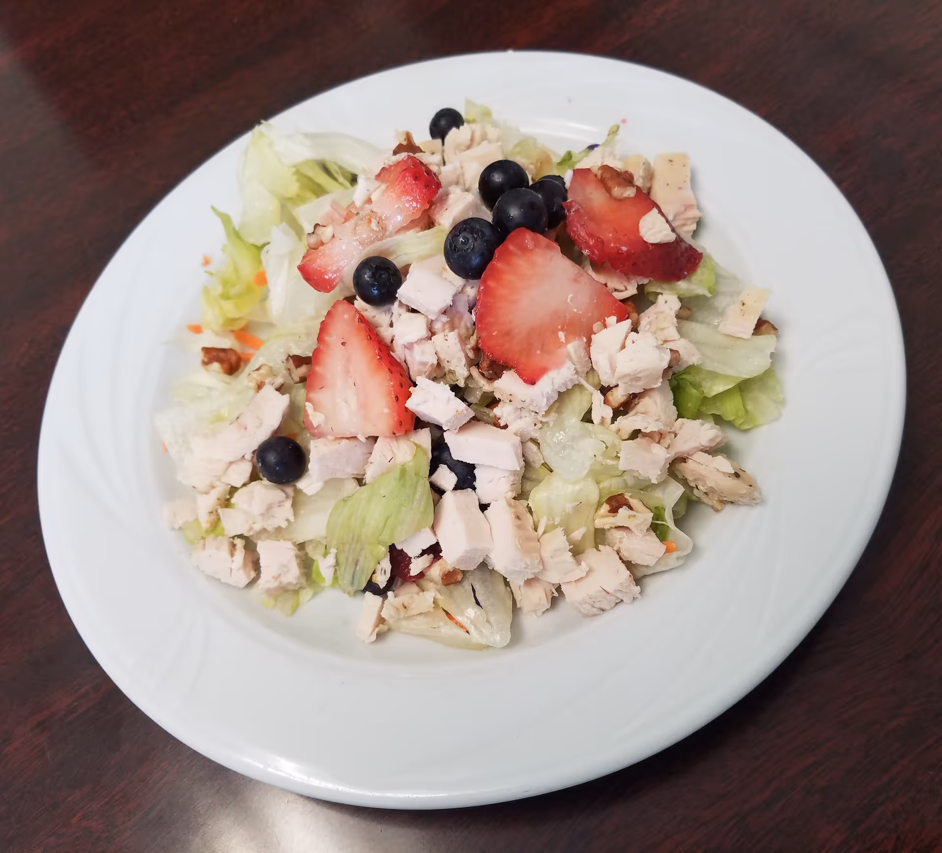 A white plate with a fresh salad consisting of chopped lettuce, diced turkey, sliced strawberries, blueberries, and small pieces of nuts, placed on a dark wooden table.