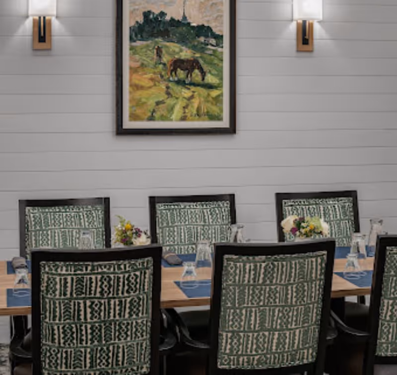 A dining room with a wooden table set with blue placemats, upside-down glasses, and small floral centerpieces. The chairs have green patterned upholstery. The wall behind is white with horizontal paneling, decorated with a framed painting of a horse in a grassy field and two wall sconces with white shades.