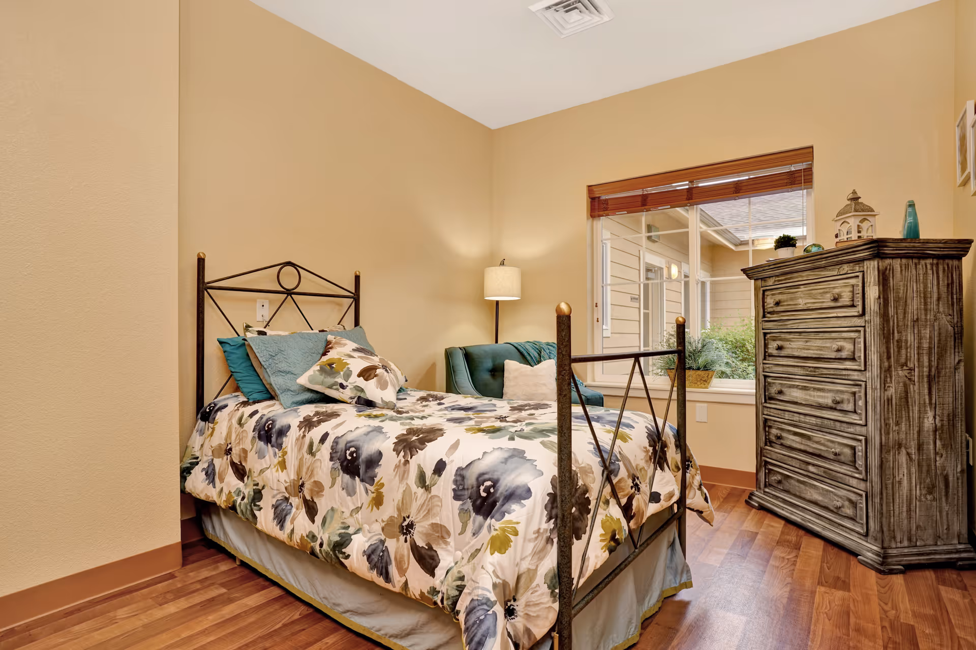A cozy bedroom with a single bed featuring a floral patterned comforter and teal pillows. Next to the bed is a teal armchair with a white cushion and a floor lamp. A large window with wooden blinds lets in natural light, and a rustic wooden dresser stands against the wall. The room has warm beige walls and wood flooring.