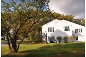 A white two-story building with green shutters surrounded by green grass and trees under a cloudy sky.