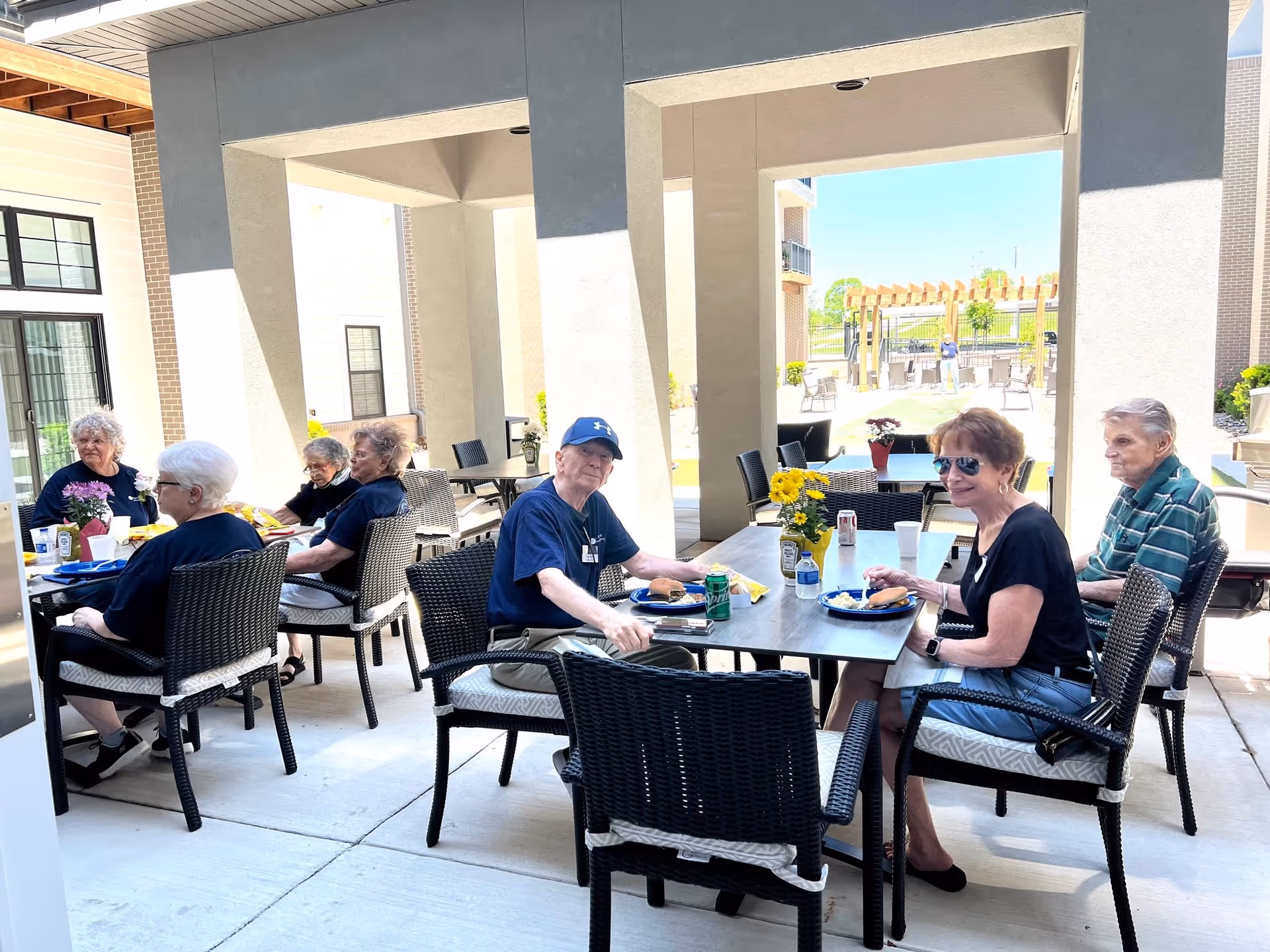 A group of elderly people sitting at outdoor tables under a covered patio, enjoying a meal together. The setting includes wicker chairs and tables with flowers and drinks, with a sunny outdoor area visible in the background.