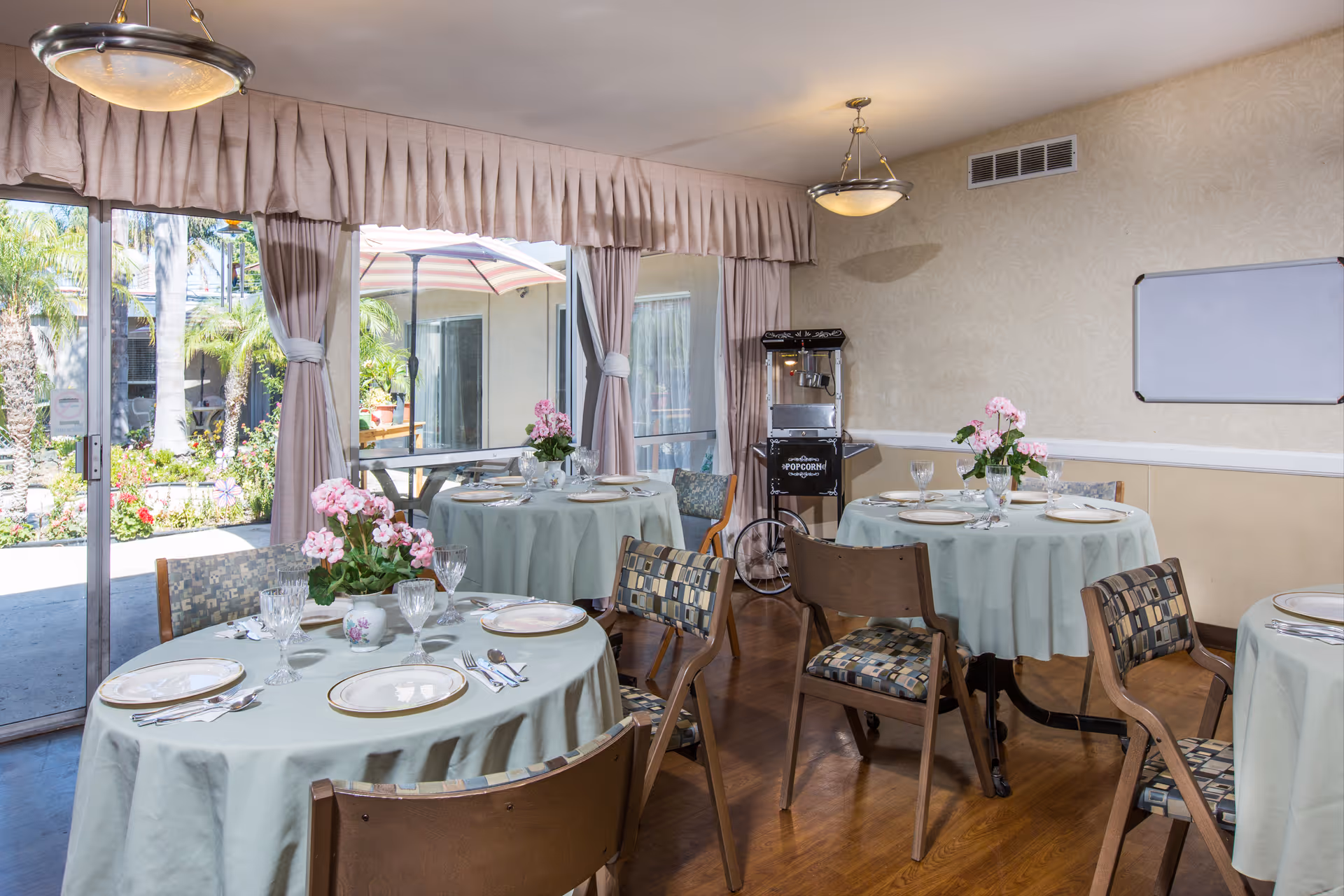 Dining room with round tables covered in light green tablecloths, set with plates, glasses, and silverware. Each table has a pink flower centerpiece. Large windows with curtains provide a view of an outdoor garden area with palm trees and flowers. A popcorn machine is visible against the wall.