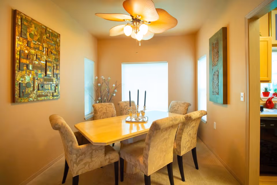 A dining room with a rectangular wooden table surrounded by six upholstered chairs with floral patterns. The room has beige walls with two pieces of wall art, three windows with blinds, and a ceiling fan with leaf-shaped blades and lights. A decorative centerpiece with three black candles is on the table.