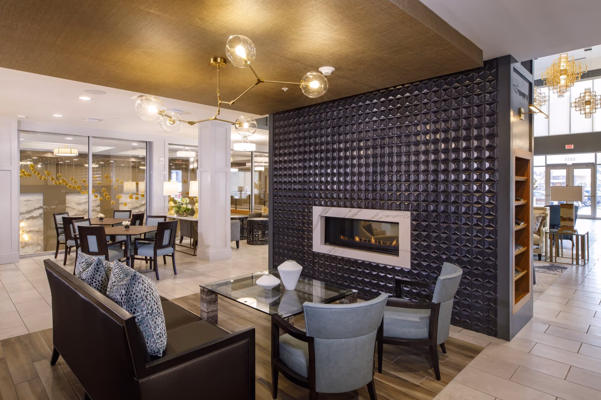 A modern and stylish common area in an assisted living facility featuring a black leather sofa with patterned pillows, two light blue armchairs, and a glass coffee table with decorative vases. The room has a textured black wall with a built-in fireplace, a gold ceiling light fixture with glass globes, and a dining area with a wooden table and chairs in the background. Large windows and glass walls allow natural light to fill the space.