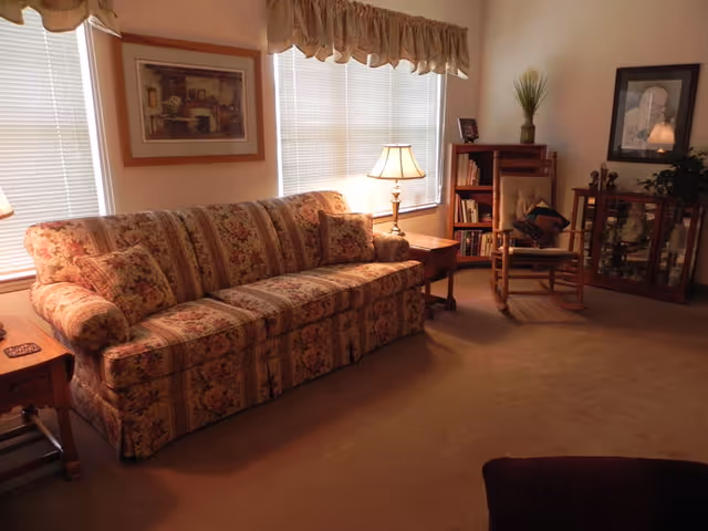 A cozy living room with a floral patterned sofa, two side tables with lamps, a rocking chair with a cushion, a bookshelf, a glass display cabinet, and framed artwork on the walls. The room has beige carpeting and windows with blinds and valances.