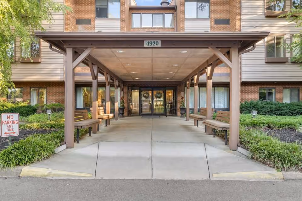 Entrance to a senior living facility with a covered walkway supported by wooden beams. There are benches on both sides under the covered area, and glass double doors lead into the building. The building exterior features brick and siding, with windows visible on the upper floors. A 'No Parking Any Time' sign is posted on the left side near some greenery.