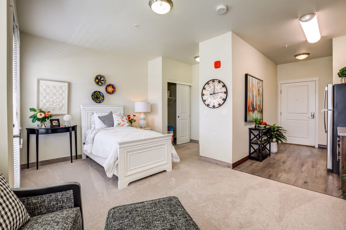 Well-lit studio apartment interior with a white bed, nightstand and lamp, seating area, and an entry/kitchen hallway.