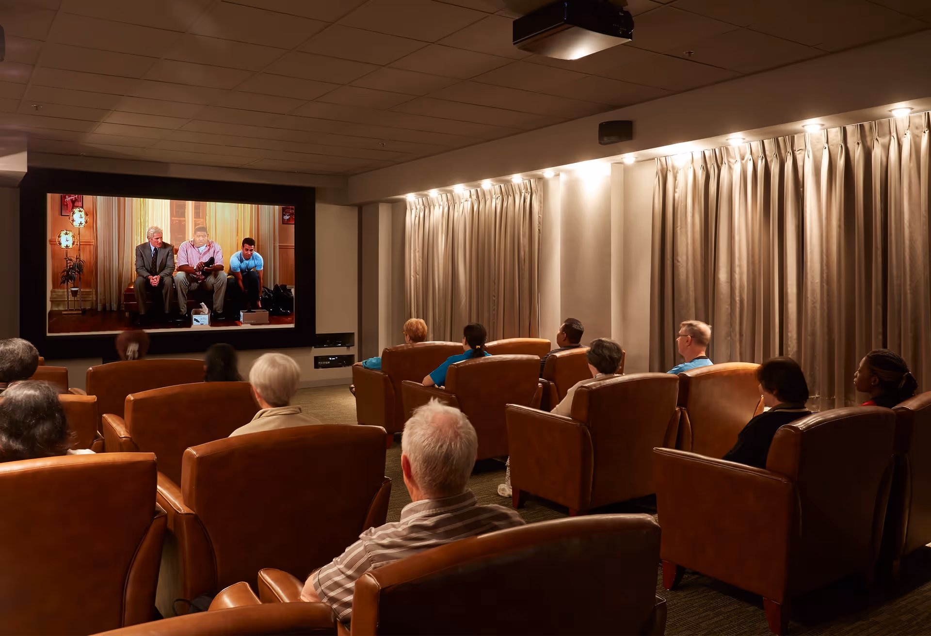 A group of elderly people seated in comfortable brown leather chairs watching a movie on a large screen in a dimly lit theater room with beige curtains along the walls.