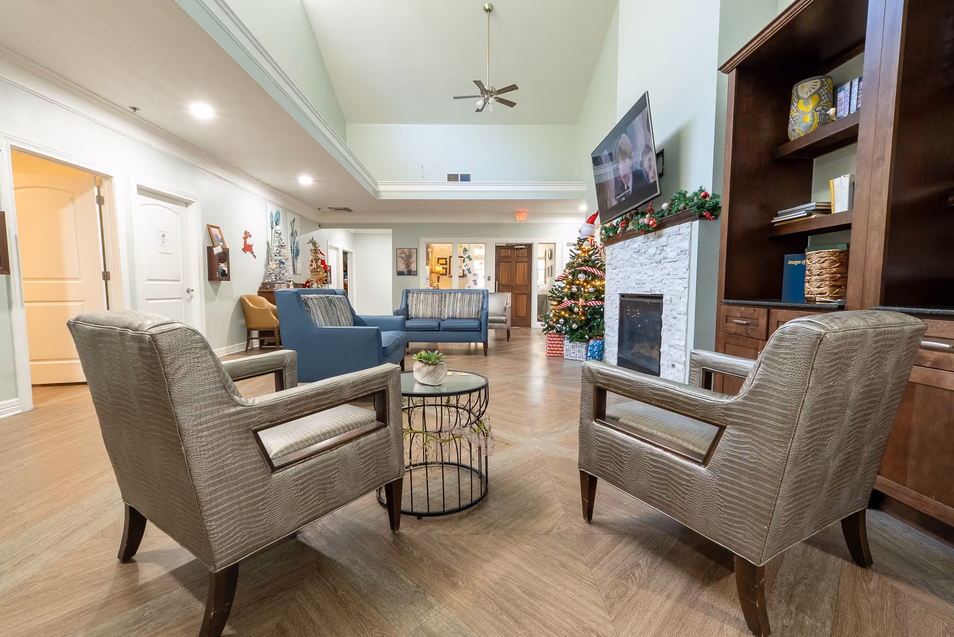 A spacious senior living facility common area with two beige armchairs facing a small round glass table, a blue sofa set in the background, a white stone fireplace with a mounted TV above it, and a decorated Christmas tree with presents underneath. The room has wooden flooring, high ceilings with a ceiling fan, and built-in wooden shelves on the right side.