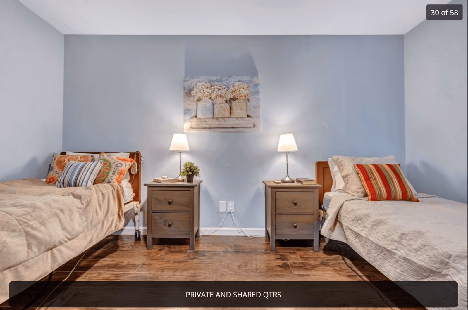 A bedroom with two single beds separated by two wooden nightstands, each with a table lamp and books. The beds have different patterned pillows and beige bedspreads. A floral painting hangs on the light blue wall above the nightstands. The floor is wooden.