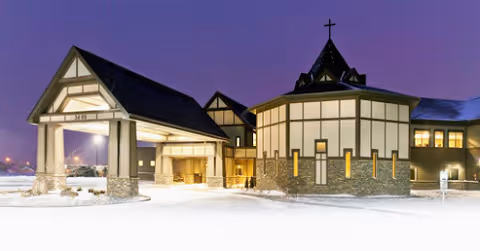 Exterior view of a large building at dusk with a covered entrance and a cross on top of a rounded section, surrounded by snow-covered ground.