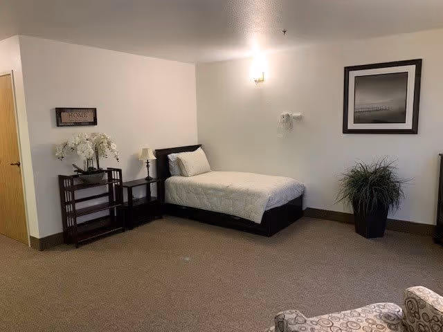 A simple bedroom with a single bed against a white wall, a small nightstand with a lamp, a decorative plant in a pot, a framed black and white photo on the wall, and a small shelving unit with white flowers. The room has beige carpet and neutral-colored walls.