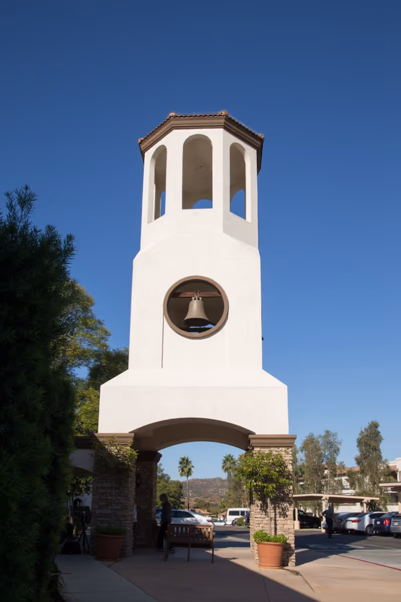 A tall white bell tower with an arched opening at the base and a bell visible in a circular opening above. The tower has a tiled roof and is surrounded by trees, potted plants, and parked cars under a clear blue sky.