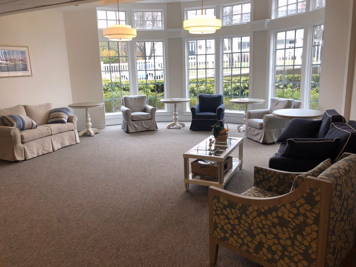 A bright and spacious living room area with large windows letting in natural light. The room features multiple armchairs and sofas in neutral and dark colors arranged around small round tables and a rectangular glass coffee table with a plant on top. The carpet is beige, and two pendant lights hang from the ceiling. Outside the windows, greenery and a white fence are visible.