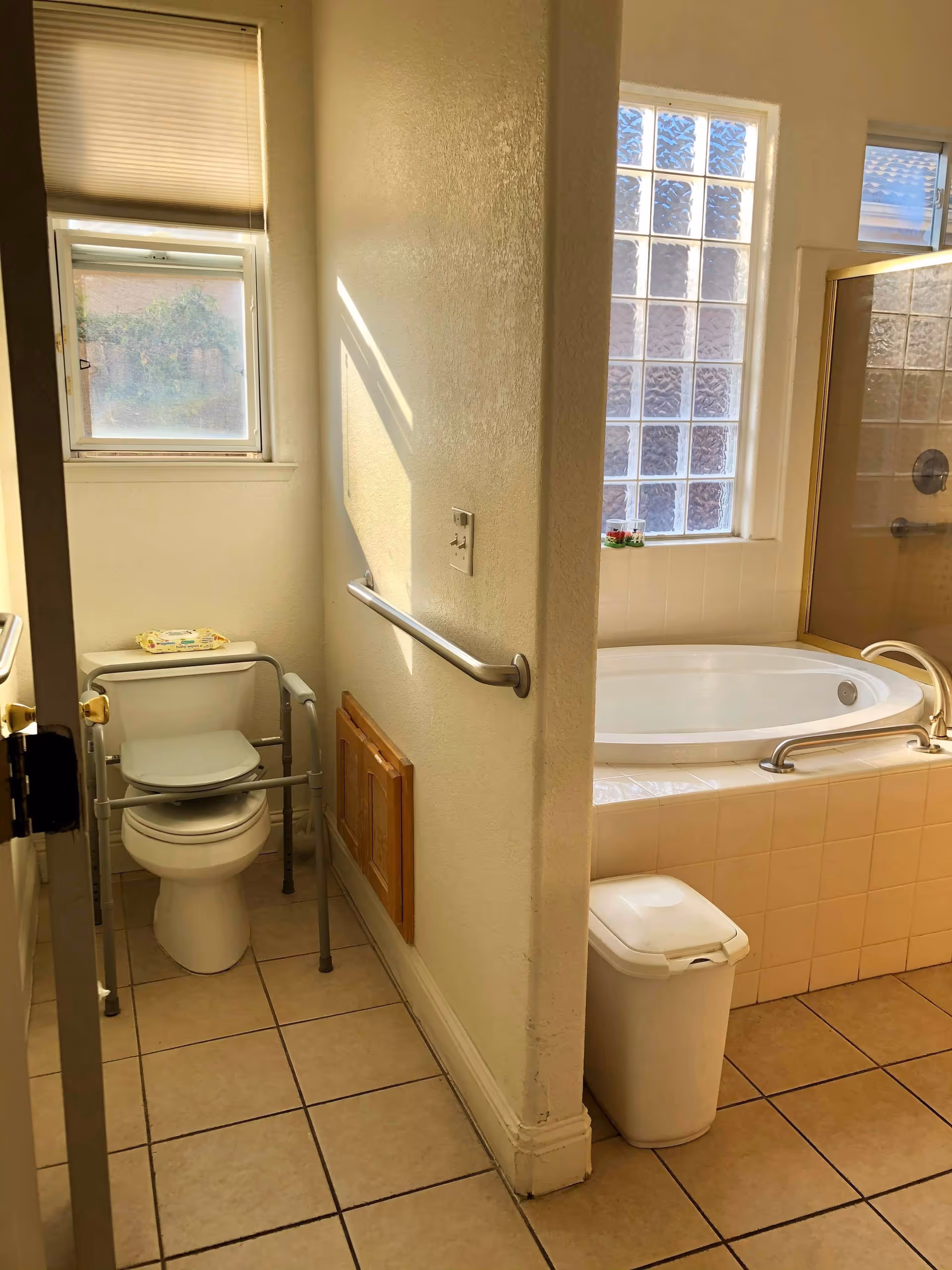 Accessible bathroom showing a toilet with a safety frame, a tiled soaking tub with grab bars, frosted windows and tiled floor.
