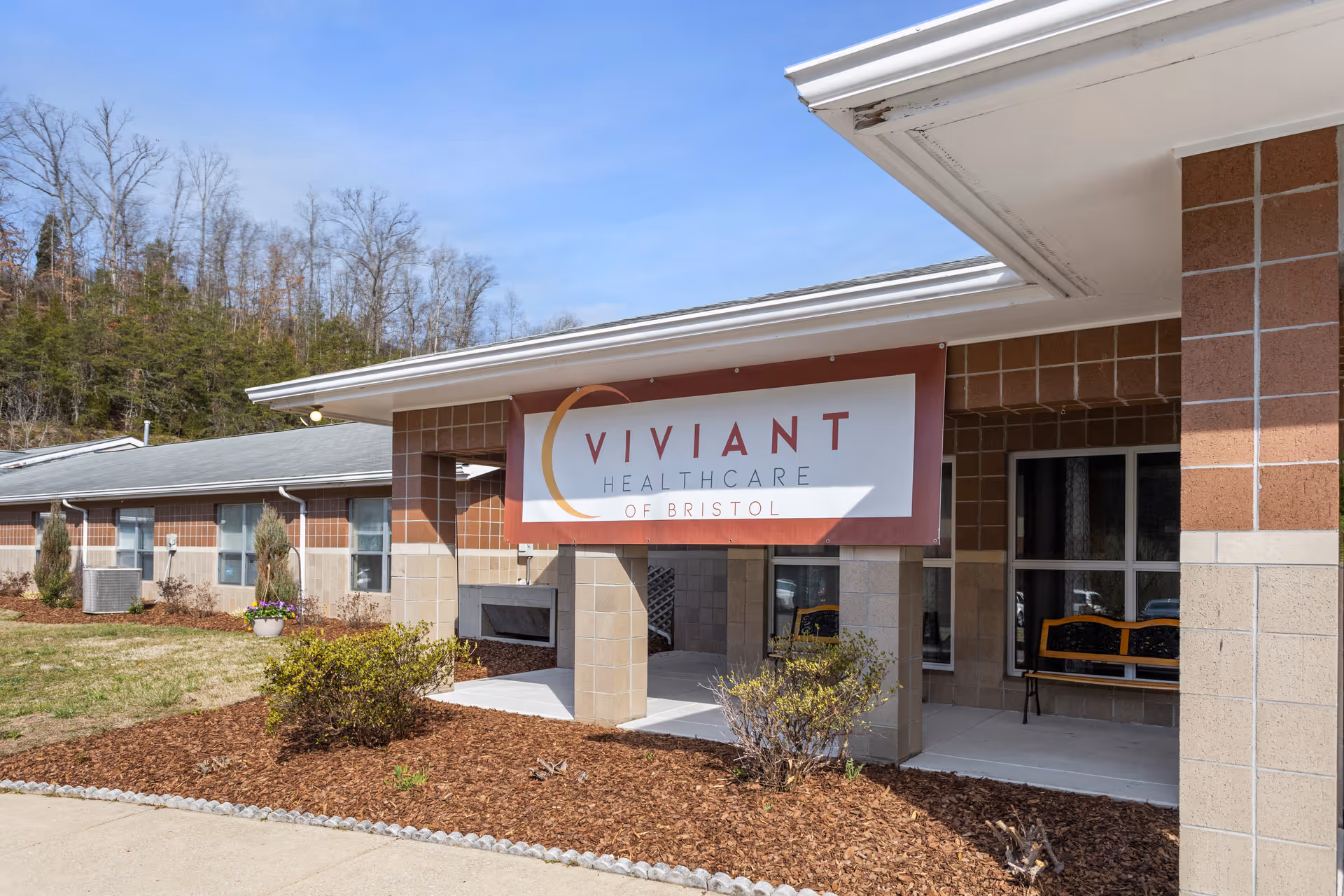 Exterior view of Viviant Healthcare of Bristol building with a covered entrance, brick and beige walls, some shrubs and plants in front, and a clear blue sky.