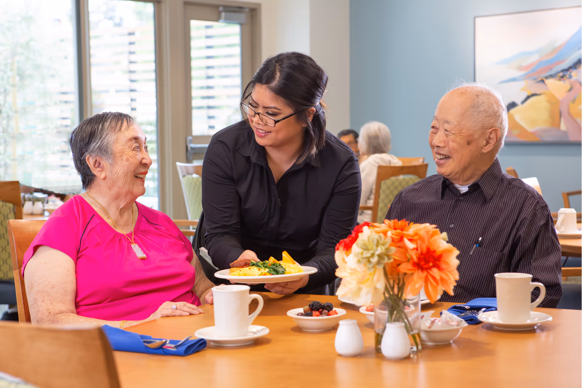 A caregiver serving a plate of food to an elderly woman in a bright pink shirt while an elderly man in a striped shirt smiles at them in a dining room setting with flowers on the table and other people in the background.