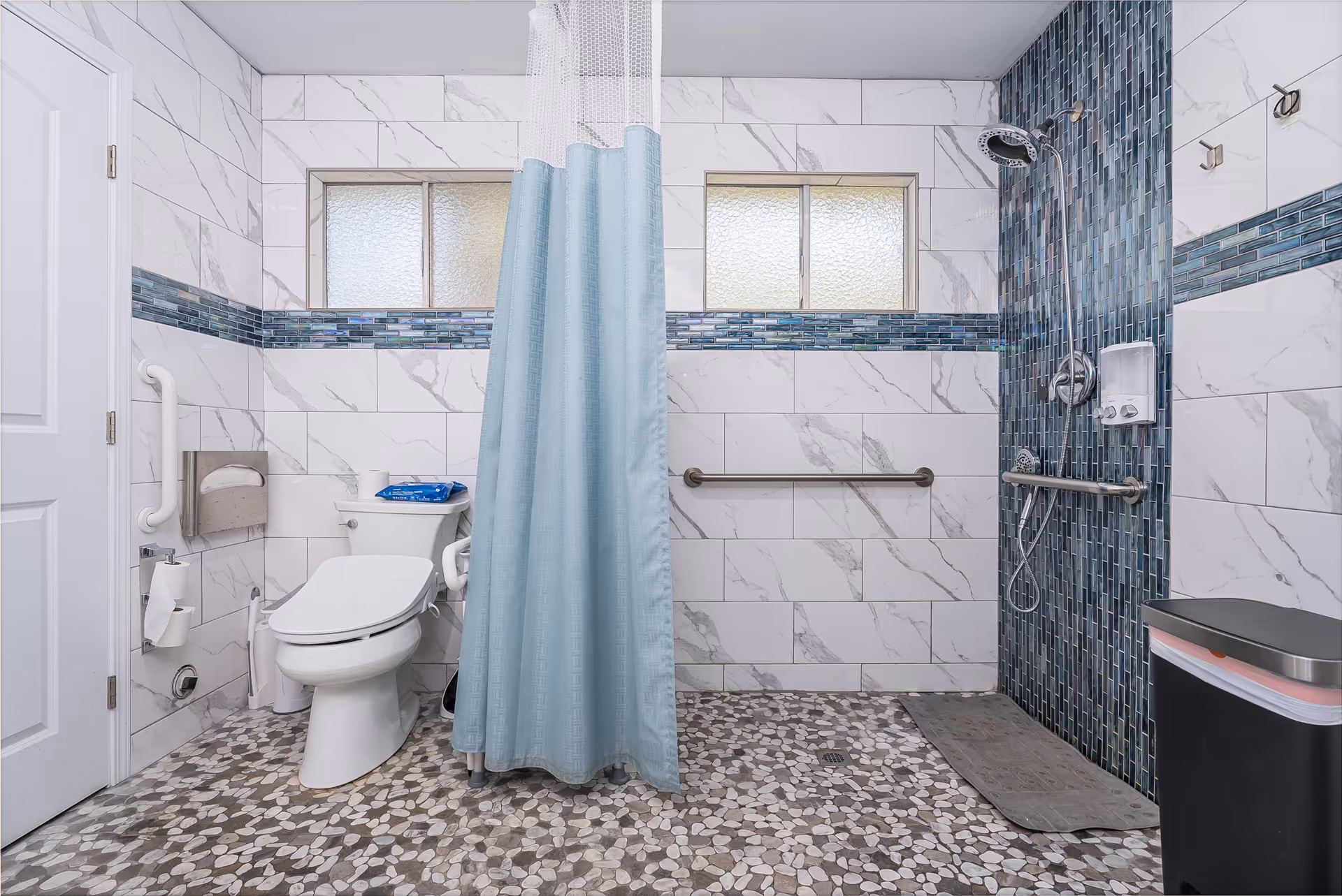 A spacious, accessible bathroom with white marble-patterned tiles on the walls and pebble-patterned flooring. The bathroom features a white toilet with grab bars, a shower area with a blue tiled accent wall, a handheld showerhead, and a blue shower curtain. There are two frosted windows above the toilet and shower area, and a trash bin is visible on the right side.